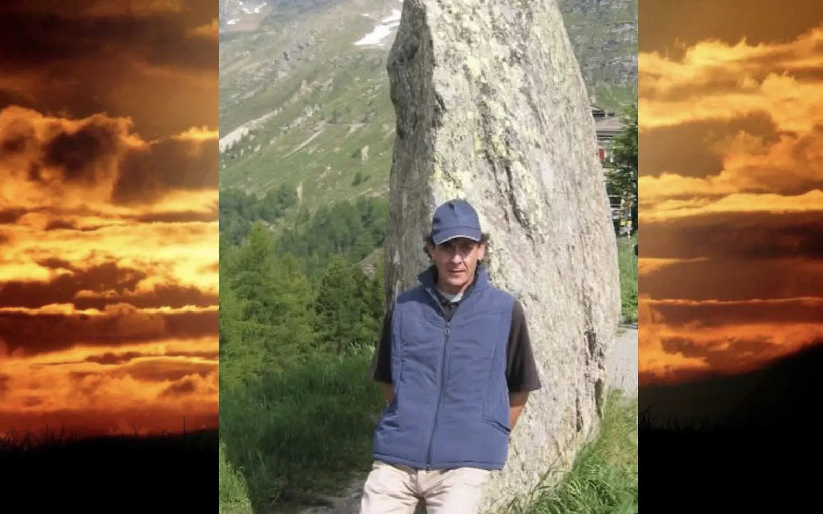 Uomo in piedi accanto a una grande roccia, con un cielo arancione e nuvoloso sullo sfondo. Indossa un gilet blu e un cappellino. Montagne verdi si vedono in lontananza.