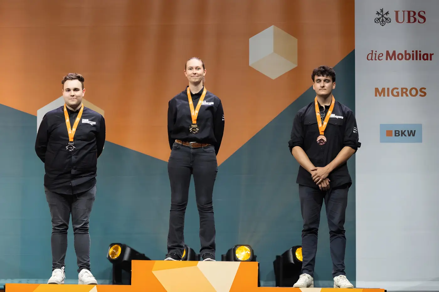 Tre vincitori in piedi su un podio, ognuno con una medaglia d'oro e una medaglia al collo. Sullo sfondo, un banner con vari loghi che include UBS e Migros.