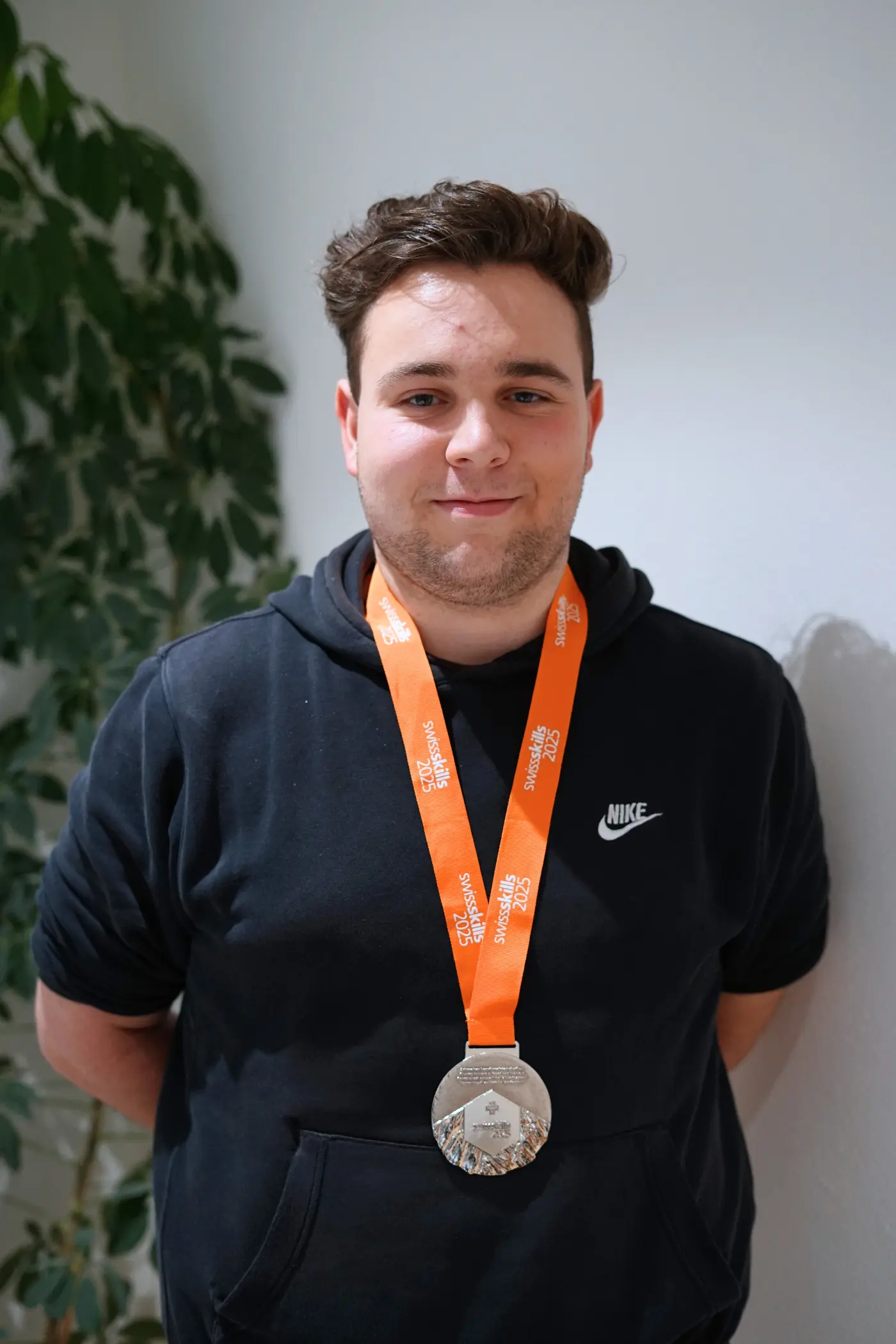 Giovane uomo sorridente che indossa una medaglia d'argento appesa a un nastro arancione. Sullo sfondo si vede una pianta.