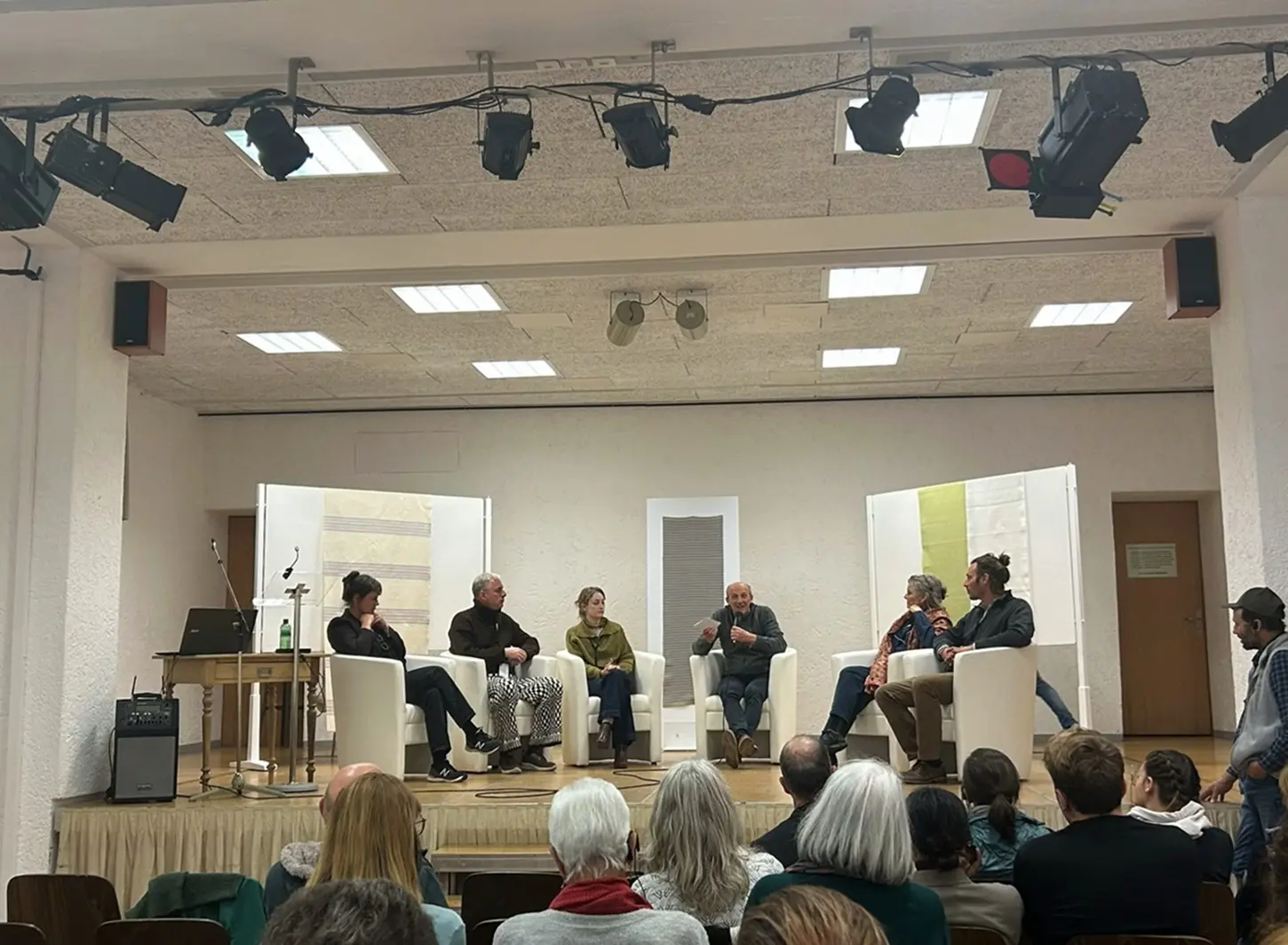 Un gruppo di sei relatori è seduto su un palco durante un evento di discussione. Sono disposti in cerchio intorno a un tavolo, con un pubblico visibile in prima fila. Lo sfondo è minimalista, con pannelli bianchi e un’illuminazione soft.