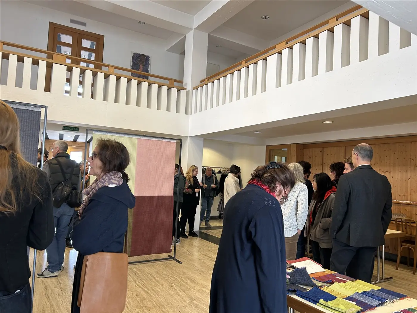 Interno di un spazio espositivo, con persone che conversano e osservano opere. Alcuni visitatori sono riuniti vicino a opere d'arte, mentre altri sono visibili sullo sfondo. La luce naturale illumina l'ambiente.