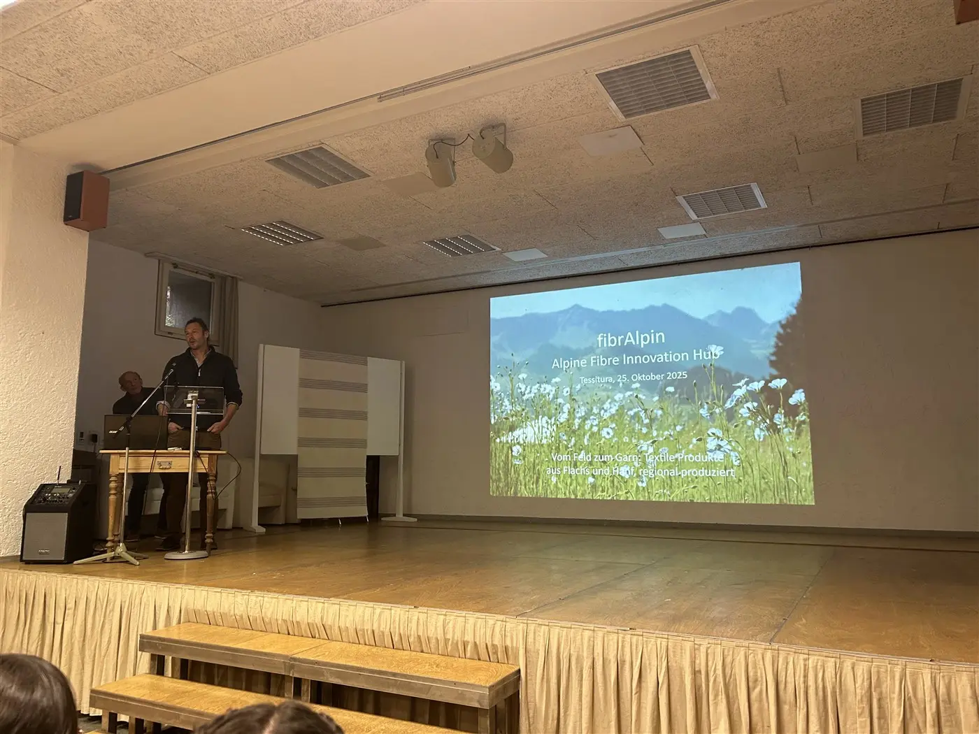 Un relatore parla di fronte a un pubblico durante un evento, con uno sfondo che mostra un paesaggio montano e un proiettore che visualizza il titolo "fib rAlpin - Alpine Fibre Innovation Hub".