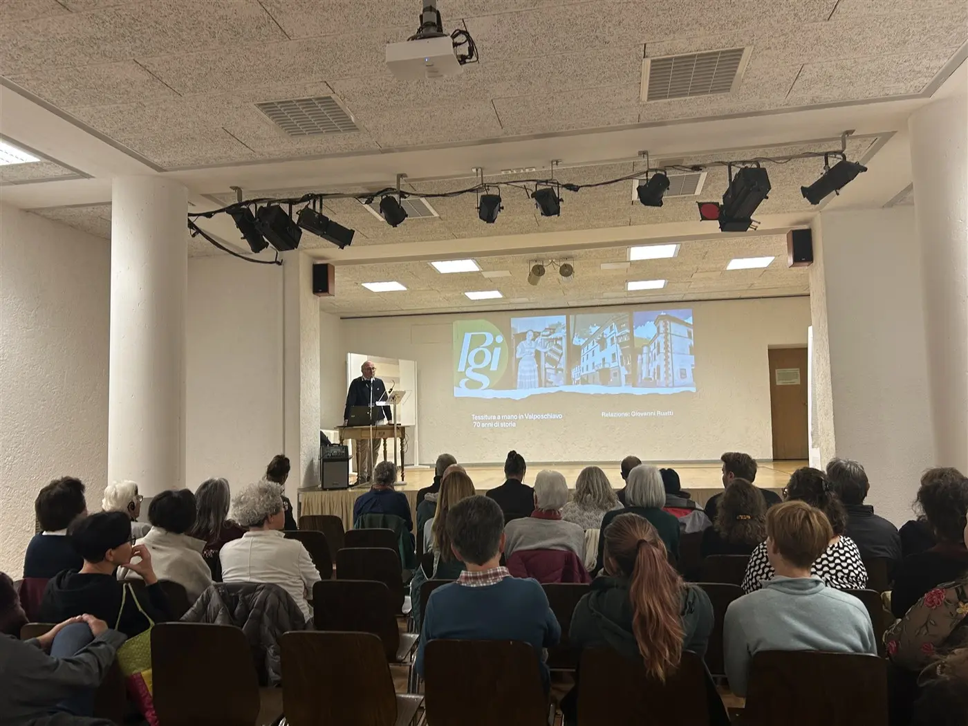 Un relatore presenta un discorso su un palco. Dietro di lui, una proiezione mostra immagini e testi. Il pubblico, composto da adulti seduti su sedie di legno, ascolta attentamente.