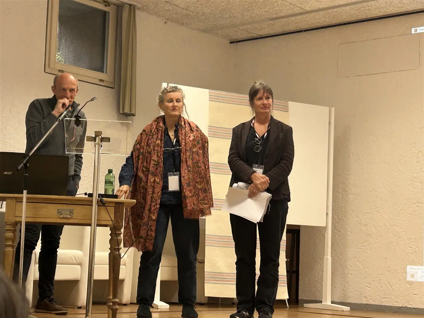 Tre relatori in un ambiente di conferenza. Un uomo con una camicia scura e una donna con un cappotto arancione sono in piedi accanto a una donna con capelli grigi e un blazer scuro. Sul tavolo ci sono un laptop e una bottiglia d'acqua. Un grande schermo è visibile sullo sfondo.