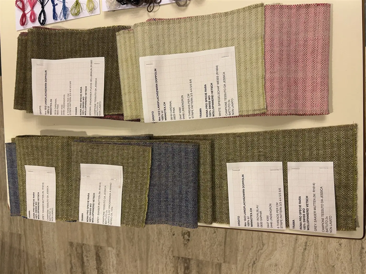 Tessuti di diverse tonalità e trame disposti su un tavolo, con etichette e informazioni accanto a ciascun campione.
