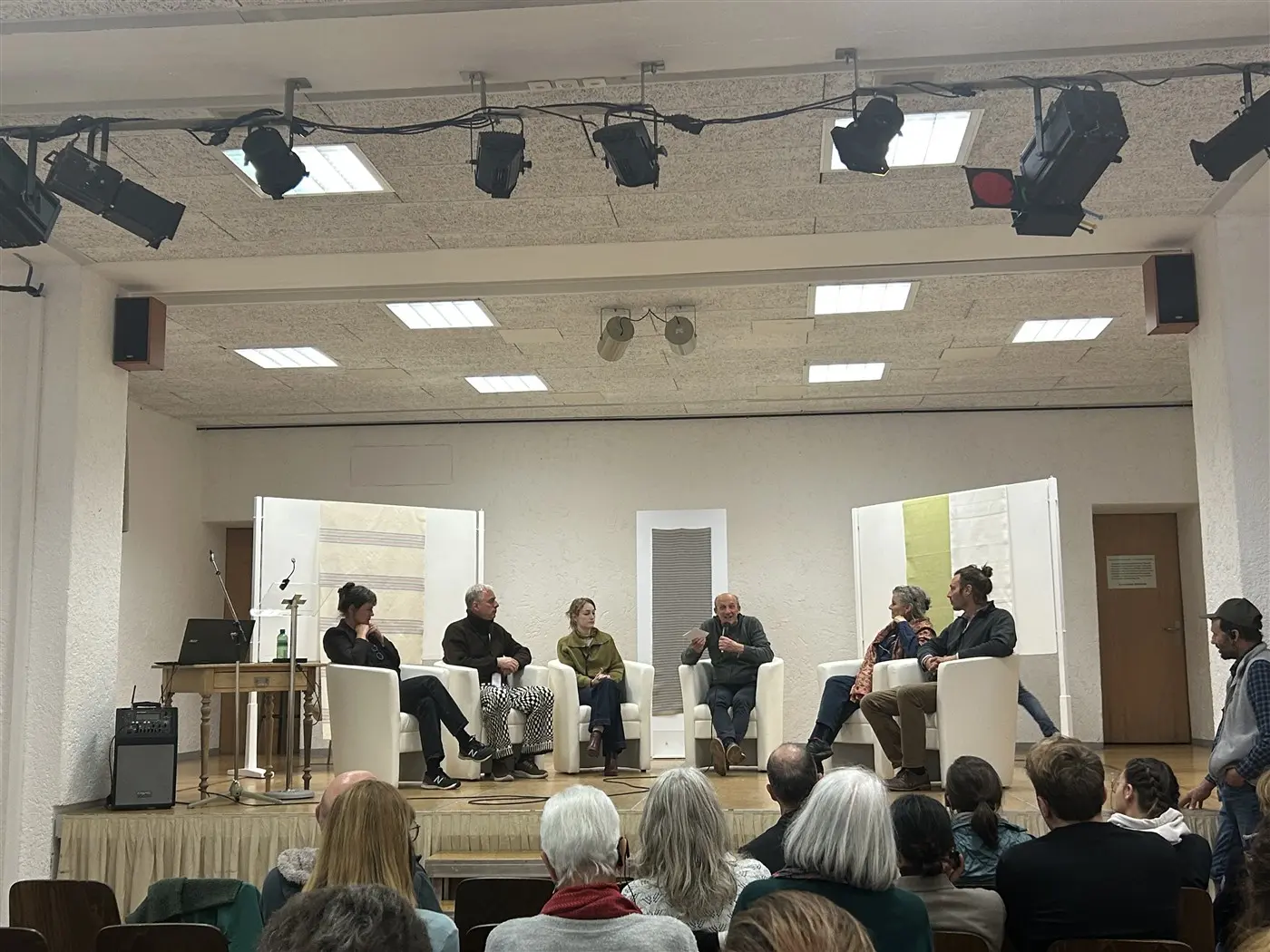 Un gruppo di cinque relatori seduti su poltrone bianche durante una discussione su un palco. Il pubblico è visibile in primo piano, mentre due grandi pannelli decorativi sono posti dietro i relatori.