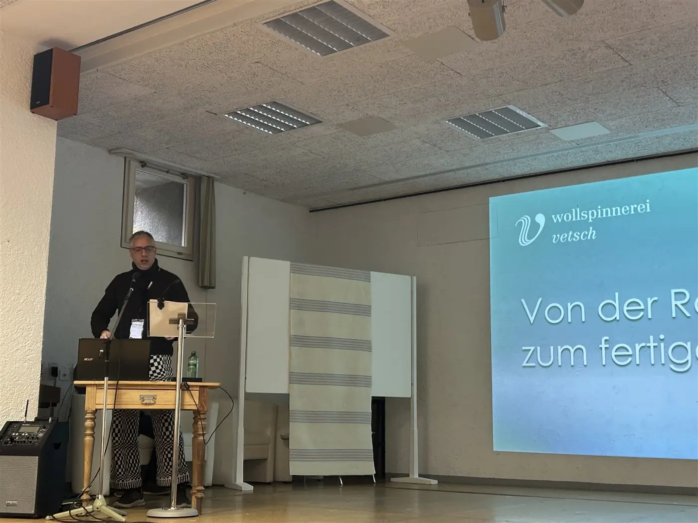 Un relatore in piedi vicino a un podio, davanti a uno schermo che mostra delle diapositive con il testo "Von der Rohwolle zum Fertigprodukt". Sullo sfondo si intravede una parete bianca e un oggetto di design.