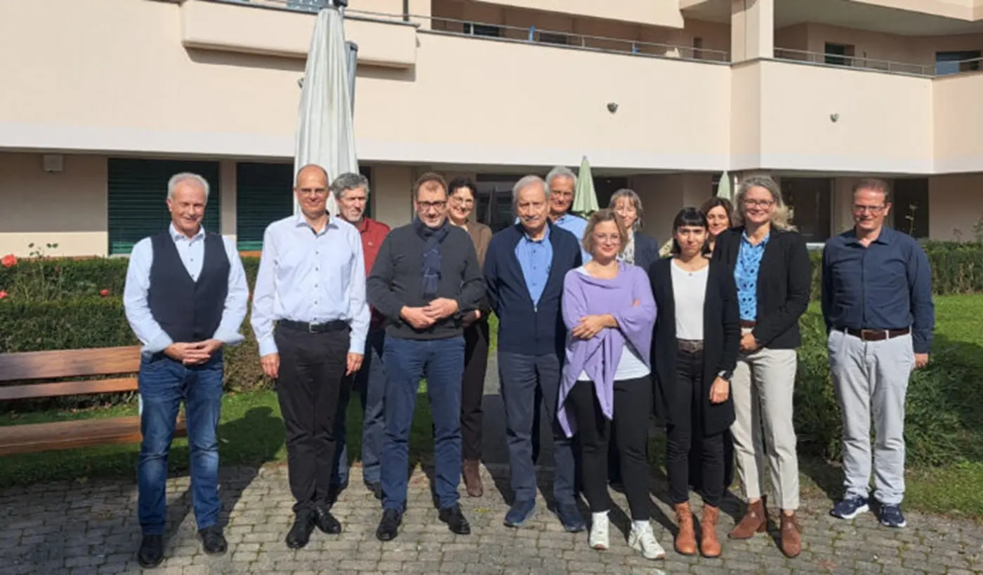 Un gruppo di persone di diverse età e stili si trova all'aperto di fronte a un edificio, sorridendo e posando per la foto. Alcuni indossano giacche e altri abbigliamenti casual. Sullo sfondo si vedono piante e una struttura architettonica.