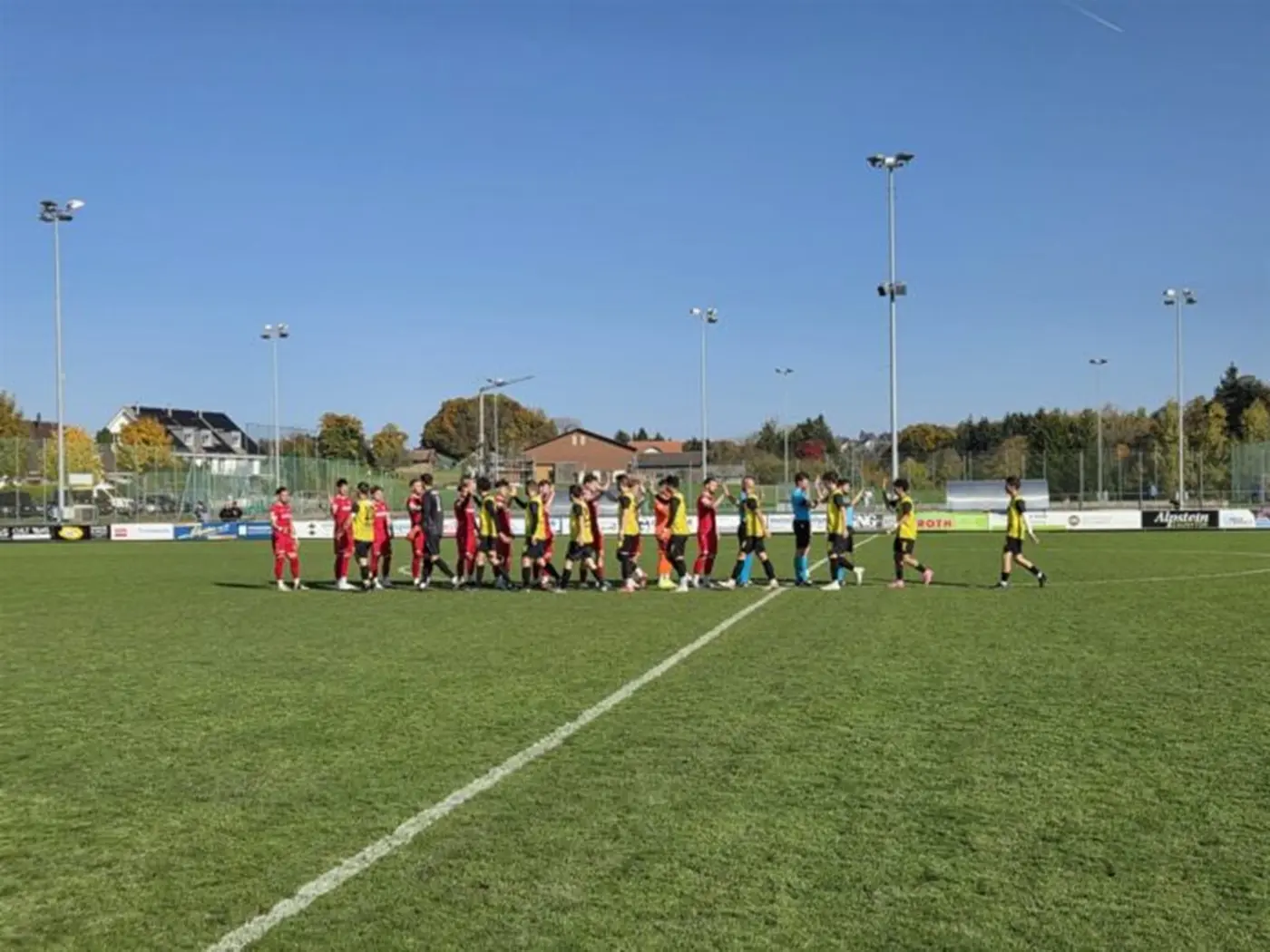 Due squadre di calcio si stringono la mano su un campo verde. Il cielo è azzurro e ci sono diversi lampioni. Sullo sfondo si vedono edifici e alberi autunnali.