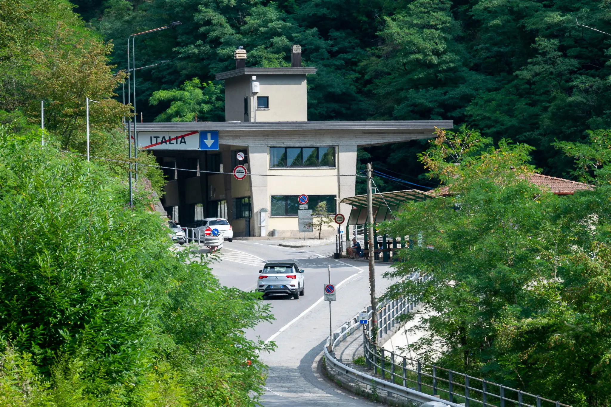 Entrata di un valico con un edificio di controllo stradale e cartelli indicativi. La strada si snoda tra la vegetazione e le montagne sullo sfondo.
