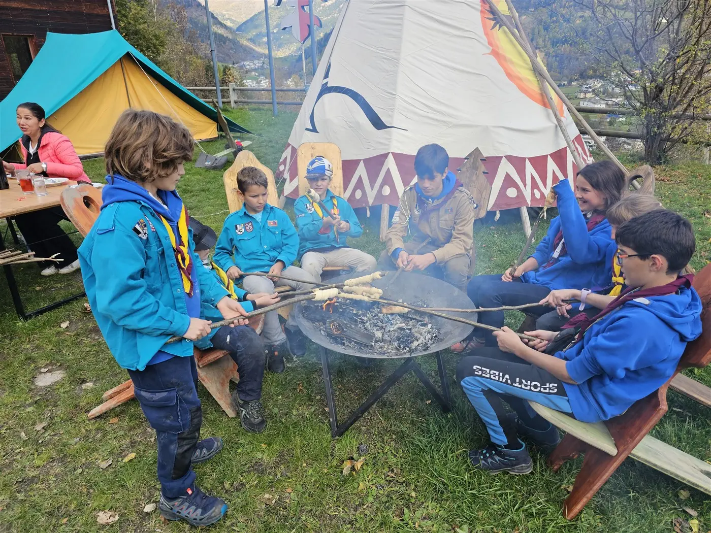 Gruppo di bambini scout seduti intorno a un falò, mentre cuociono delle pannocchie su bastoncini. In lontananza si vedono tende colorate e alberi. Ambiente naturale con atmosfera di attività all'aperto.