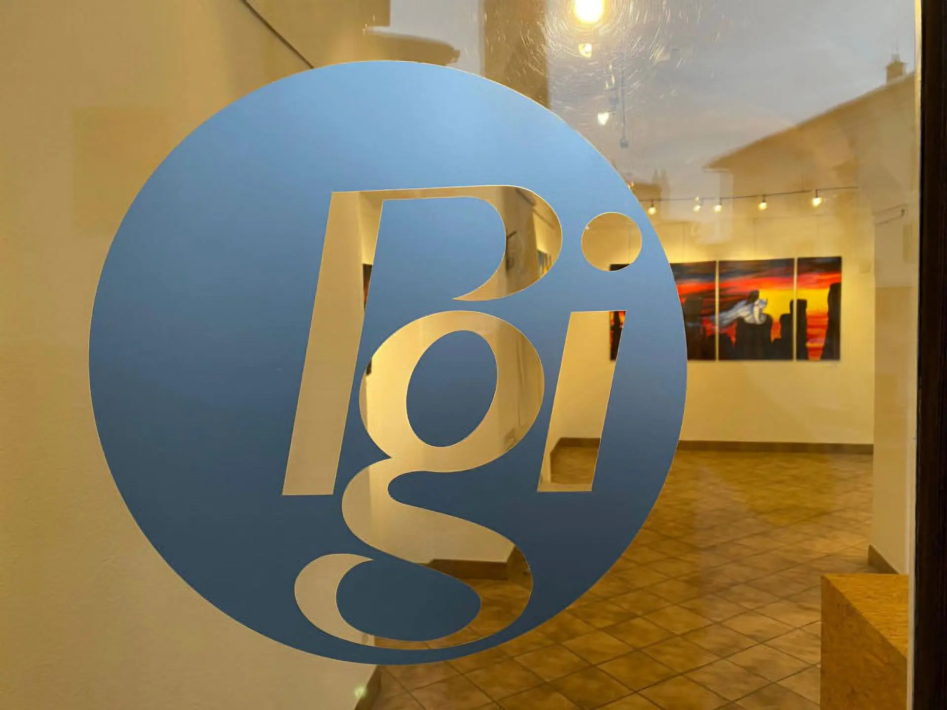 Logo blu con le lettere "Pgi" su uno sfondo chiaro, visibile attraverso una finestra di un'esposizione artistica. Alle pareti si notano opere d'arte appese.