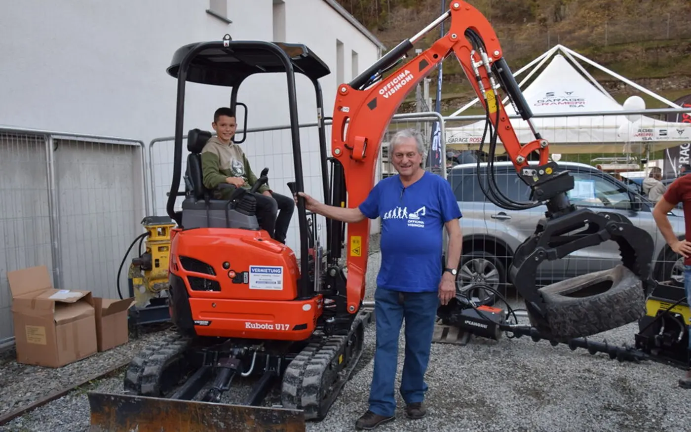 Un uomo anziano sorride accanto a un ragazzo seduto su un mini escavatore arancione. Sullo sfondo si vedono vari attrezzi e un veicolo.