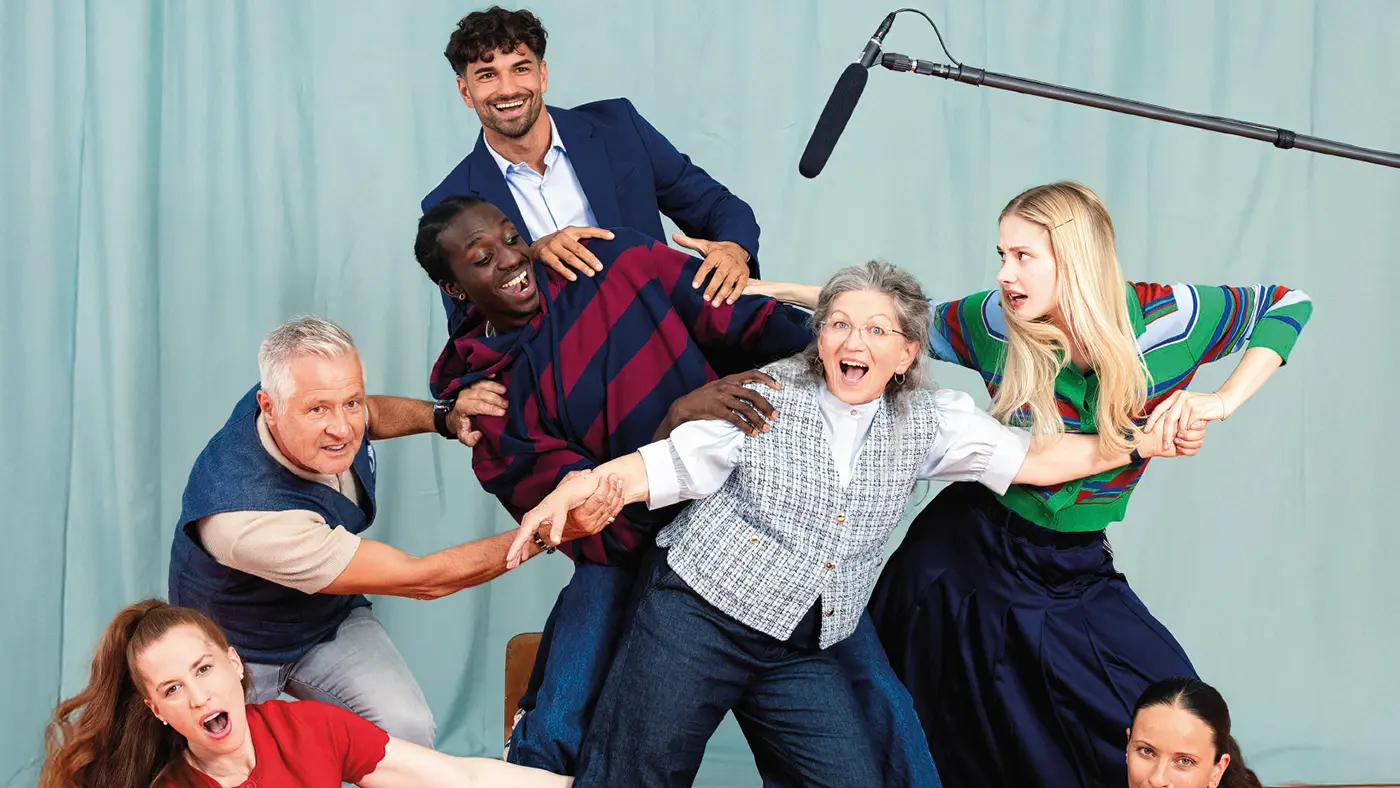 Un gruppo di persone di diverse età e origini si trova in una posa scenica divertente, con espressioni esagerate e movimenti dinamici. Alcuni si abbracciano e si afferrano, mentre uno tiene un microfono sopra di loro. Sullo sfondo, un drappo colorato.