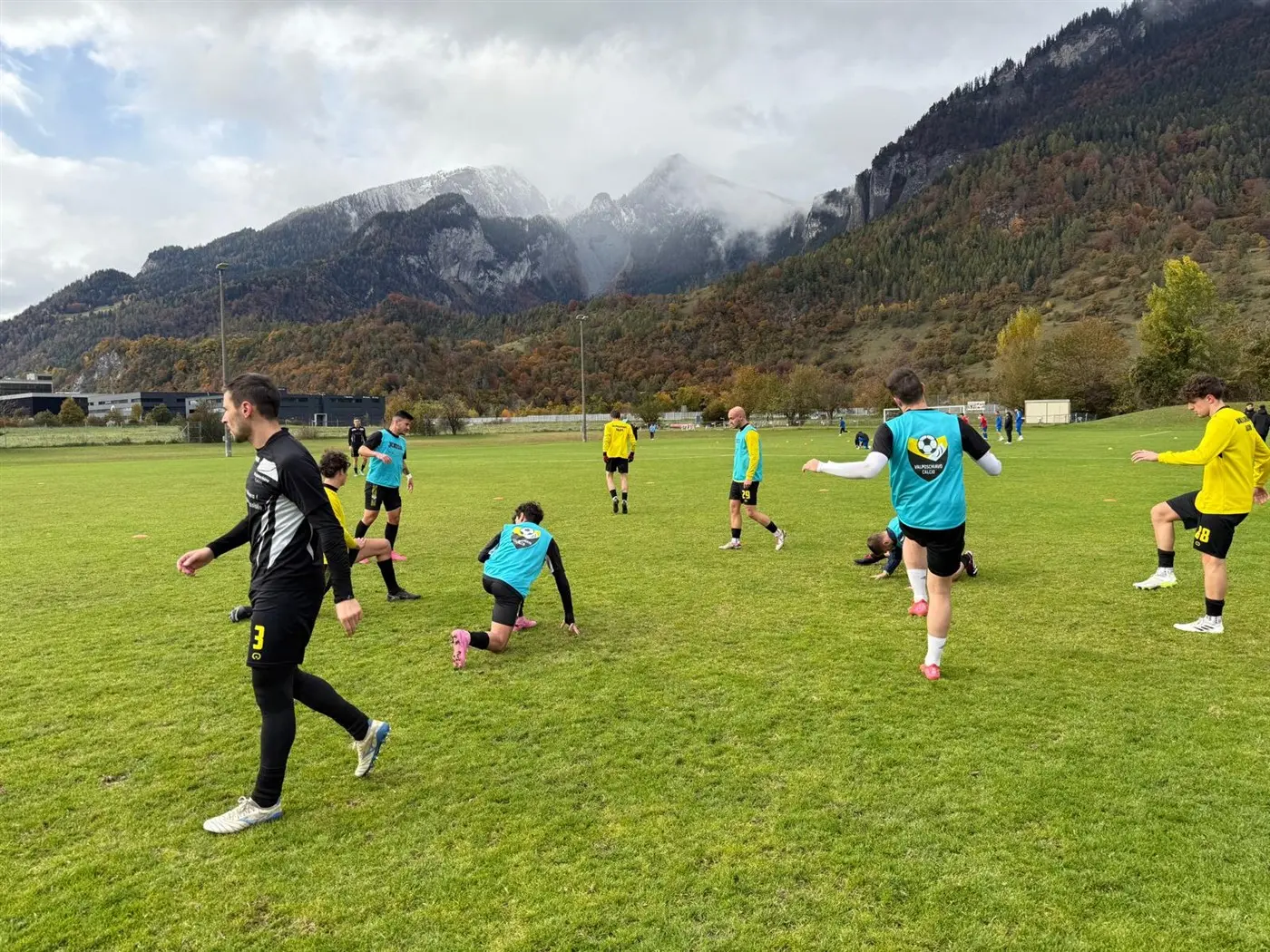 Giocatori di calcio si allenano su un campo verde, con montagne sullo sfondo. Alcuni si allungano mentre altri si muovono attivamente. Il cielo è nuvoloso con segni di pioggia.