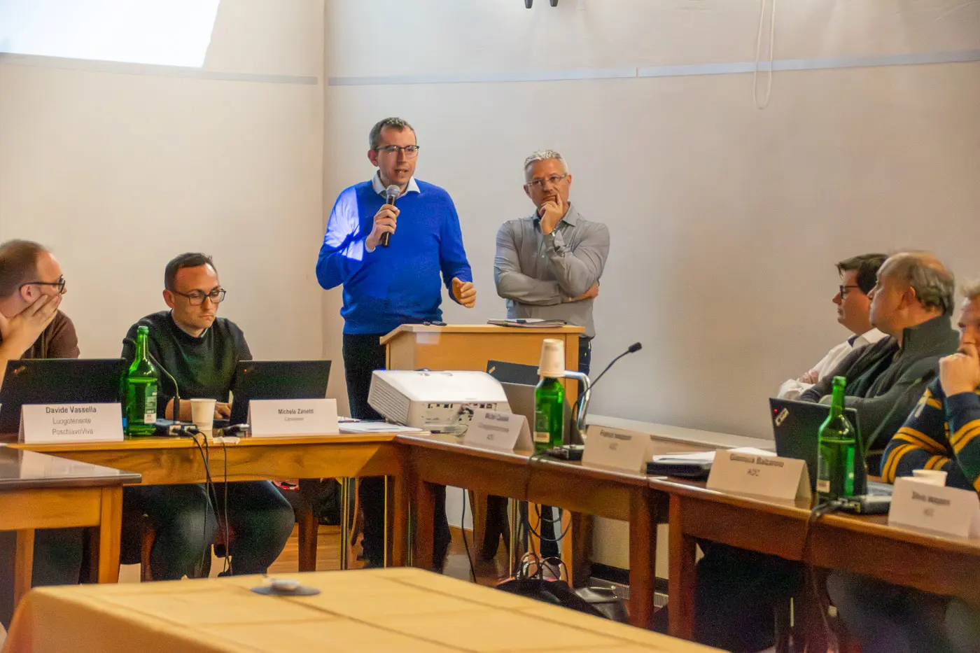Due uomini parlano a un tavolo di conferenza. Un uomo in maglione blu usa un microfono, mentre l'altro, con occhiali e maglione grigio, ascolta. Altri partecipanti sono seduti attorno al tavolo con laptop e bottiglie d'acqua.