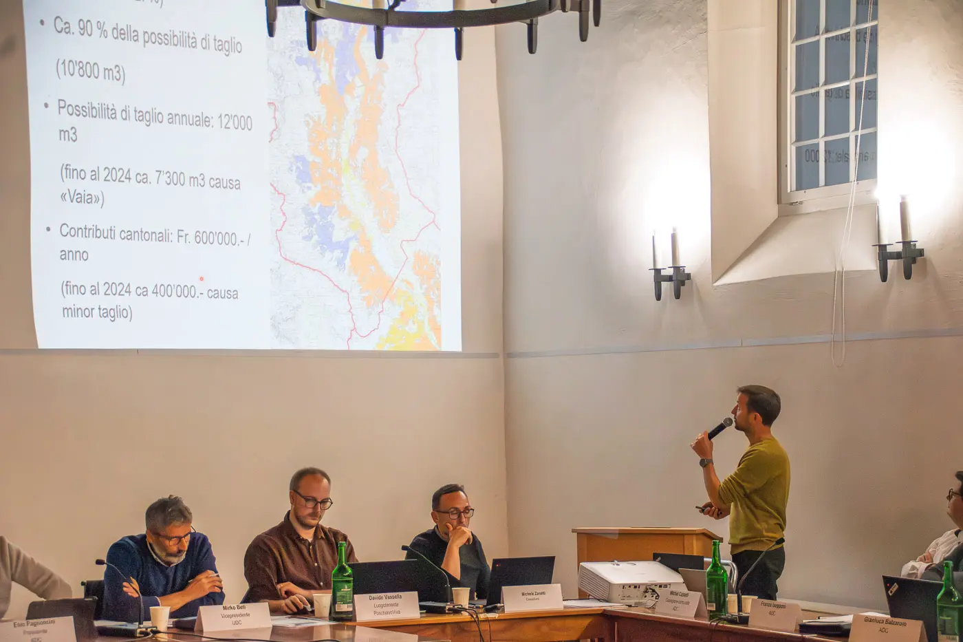 Un uomo parla a un tavolo di relatori durante una presentazione, con mappe e grafici sullo sfondo. I relatori, composti da uomini in abiti formali, ascoltano attentamente.