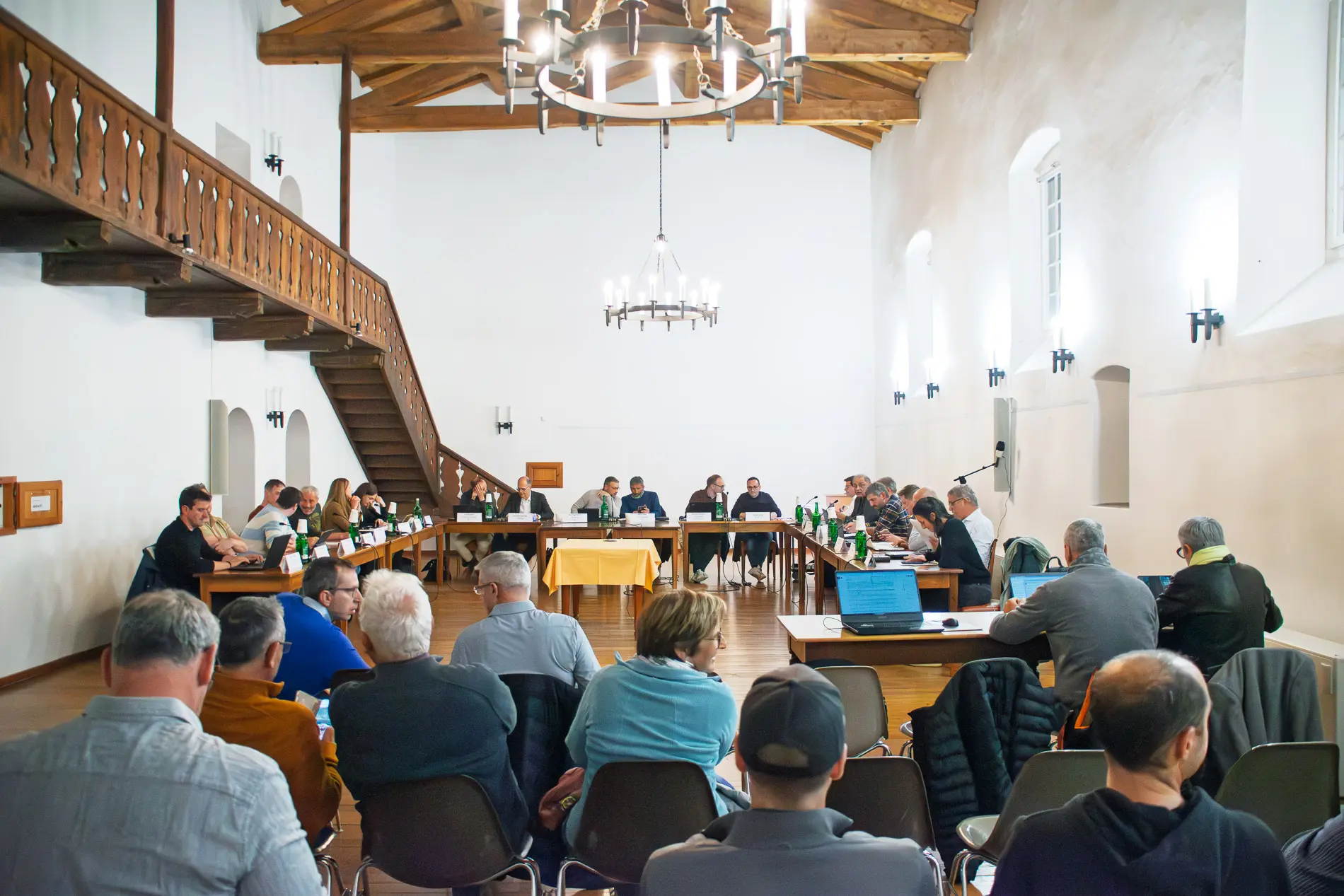 Un gruppo di persone sedute attorno a tavoli in una sala riunioni con soffitto in legno. Alcuni partecipanti sono rivolti verso un gruppo di relatori, mentre altri osservano. La sala è ben illuminata con lampadari e finestre.