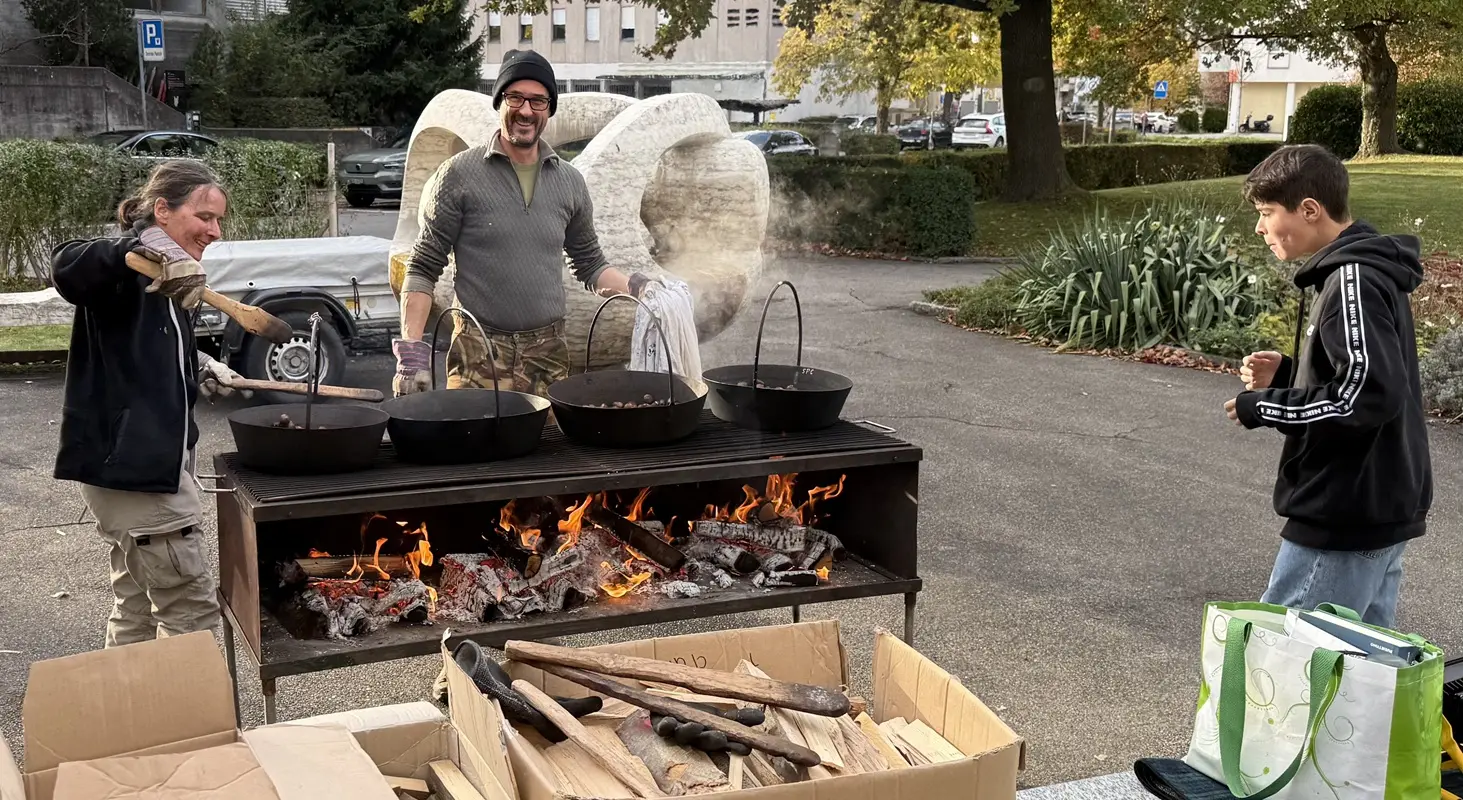 Tre persone stanno cucinando all'aperto su un grande barbecue. Due adulti, uno con capelli lunghi e l'altro con occhiali, mescolano contenitori neri sopra delle braci ardenti. Un giovane osserva mentre si prepara a cucinare. Sullo sfondo, ci sono sculture e un'area verde.