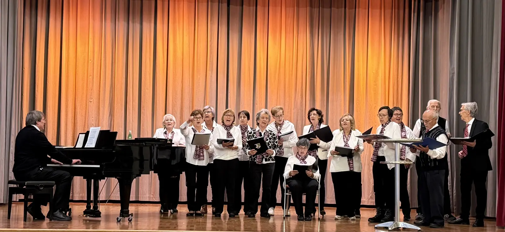 Un coro di persone anziane canta su un palcoscenico, con un pianista che suona al pianoforte. Indossano camicie bianche e sciarpe scure. Sullo sfondo, una tenda arancione illumina l'evento.