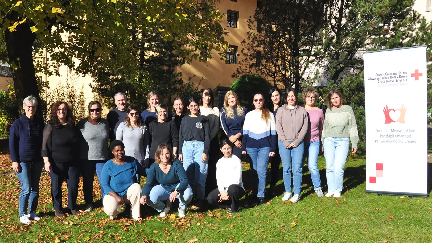 Gruppo di persone sorridenti posizionati all'aperto, in un parco con alberi autunnali. Si trovano davanti a un banner del Croce Rossa Italiana.