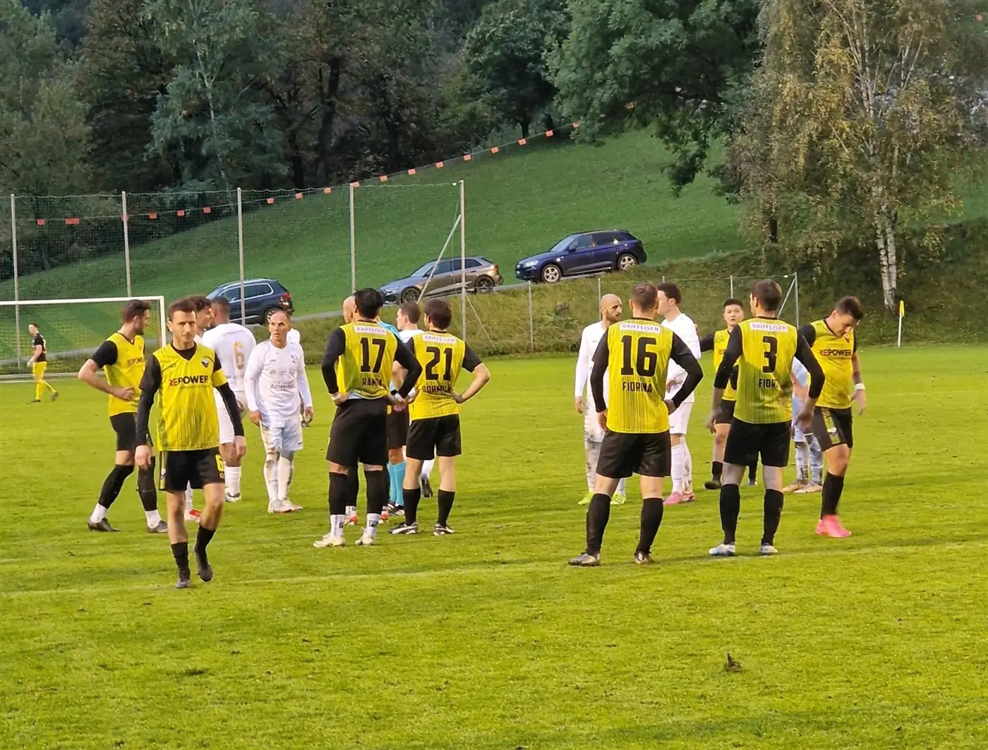 Giocatori di calcio in campo durante una pausa, indossando maglie gialle e nere. Alcuni si trovano in cerchio e discutono tra loro, mentre altri sono in secondo piano. Sullo sfondo si vedono auto parcheggiate e alberi.
