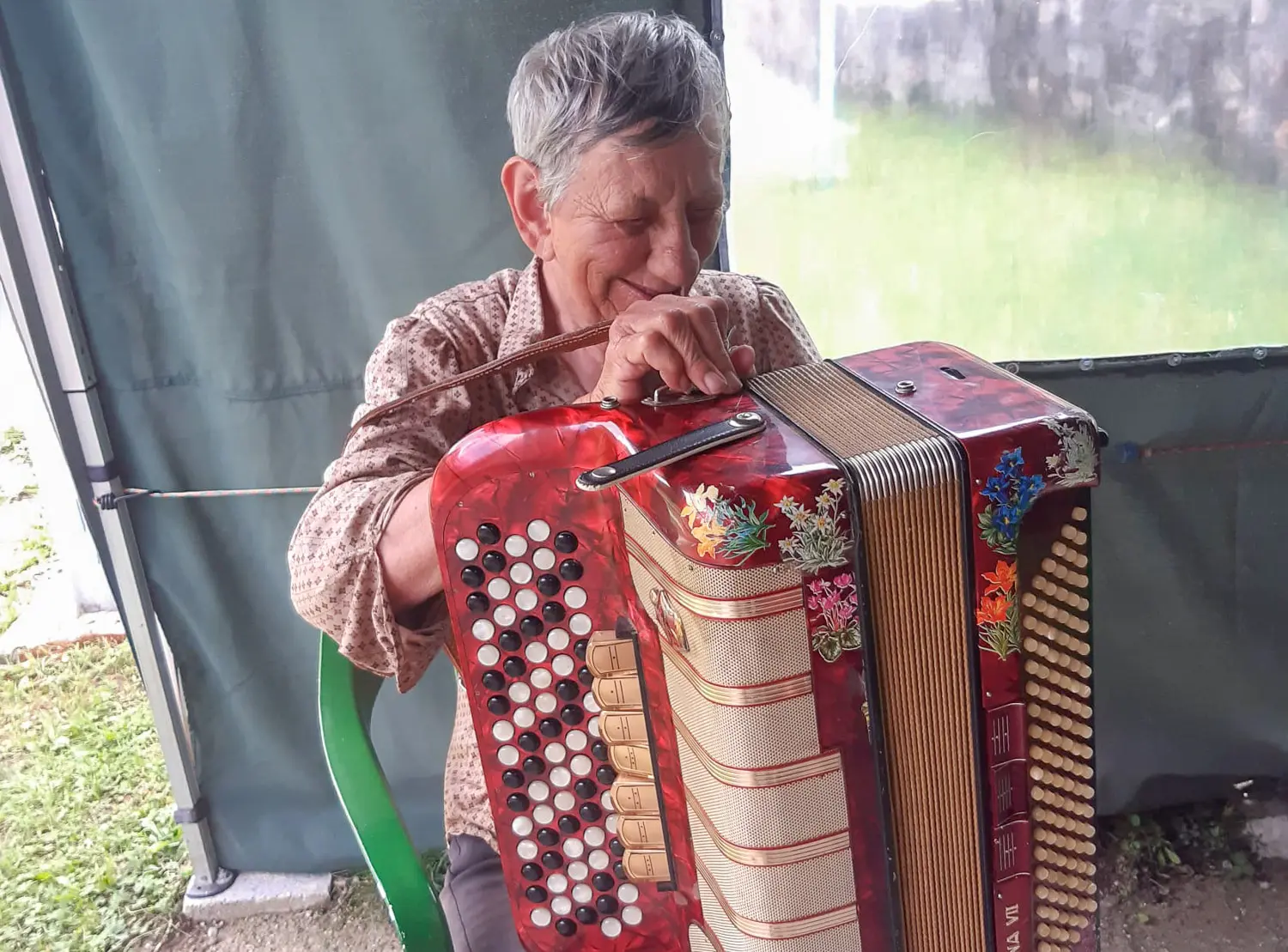 Una donna anziana suona un'accordion rossa mentre sorride, seduta su una sedia verde in un ambiente all'aperto.