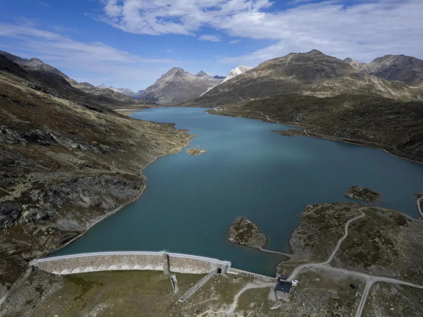 Vista panoramica di un lago blu circondato da montagne, con una diga in primo piano. Il cielo è prevalentemente nuvoloso, creando un'atmosfera tranquilla e serena.