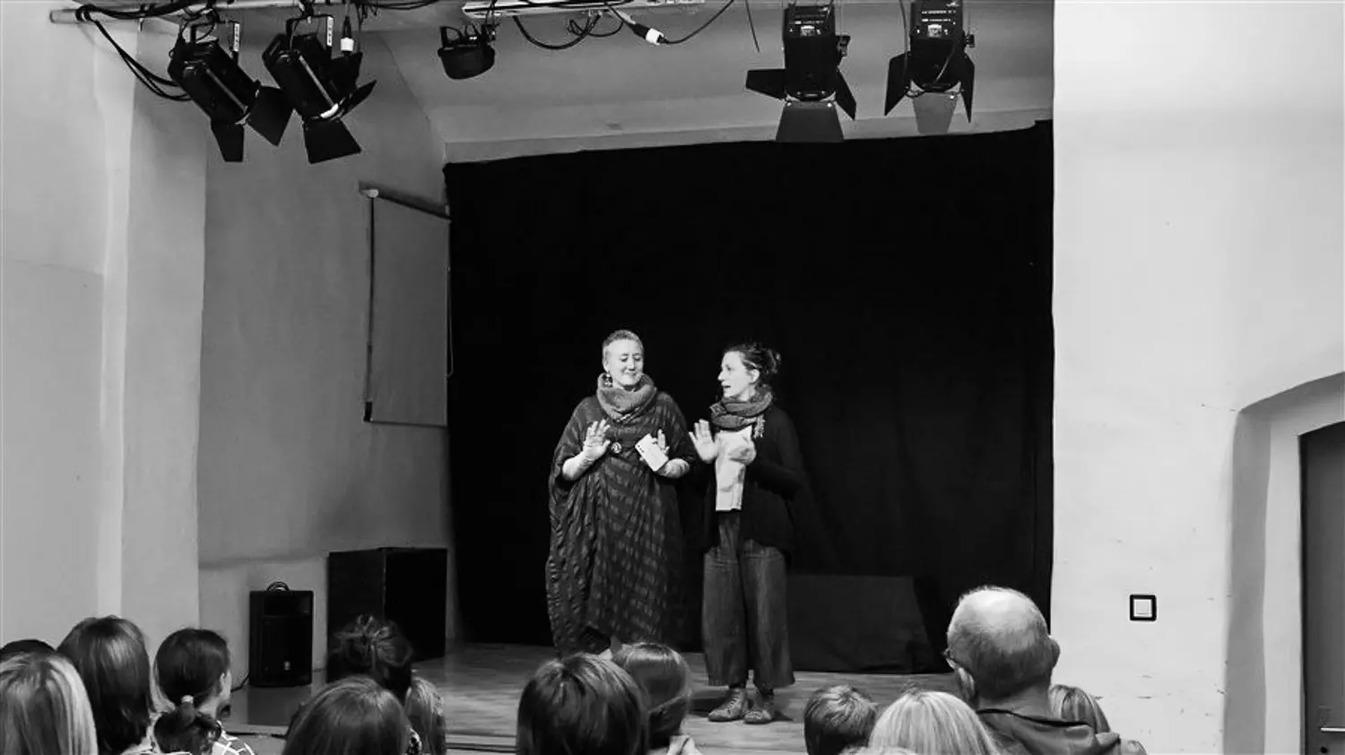 Due donne sul palcoscenico di un teatro, sorridendo e interagendo con il pubblico. Sullo sfondo, un drappo nero e luci sopra di loro. Davanti, un pubblico attento e curioso.