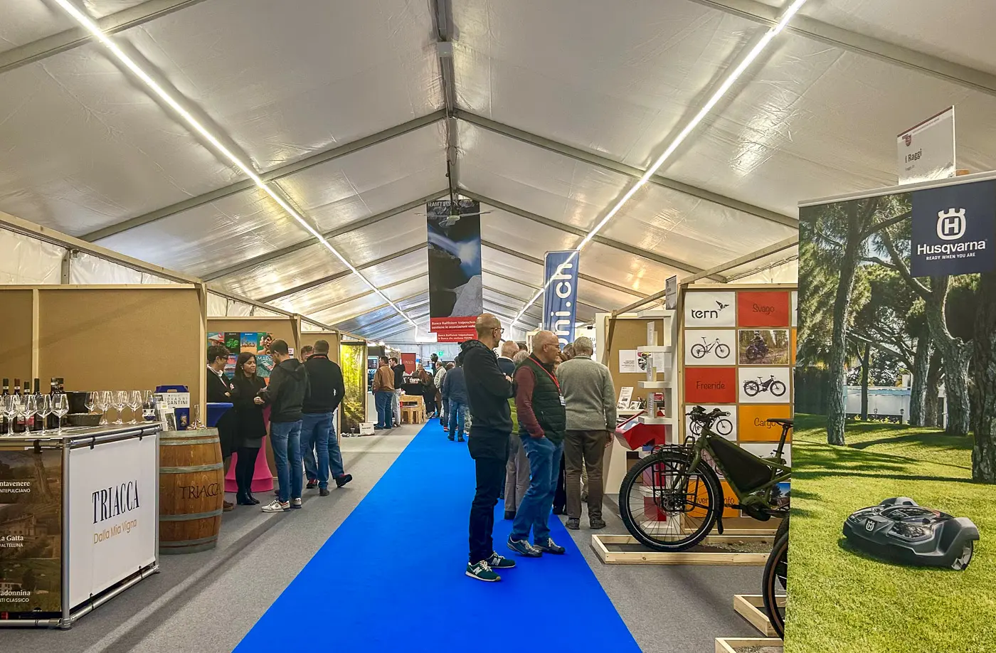 Interno di una fiera allestita in una tenda, con stand che espongono biciclette e prodotti, oltre a persone che girano tra i vari espositori. Un tappeto blu percorre il corridoio centrale.