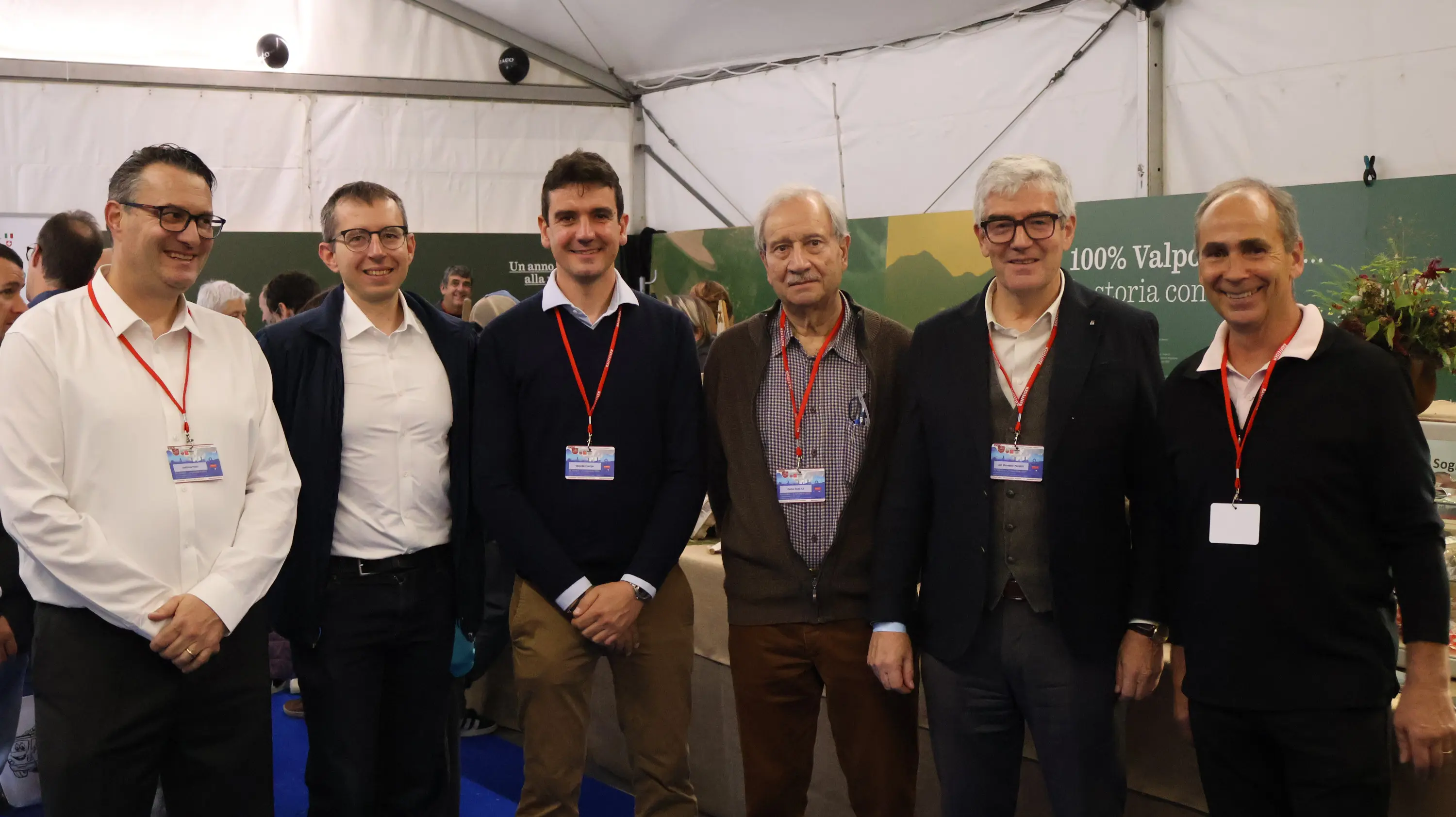 Sei uomini sorridenti si trovano all'interno di una tenda per un evento. Indossano abiti formali e hanno distintivi identificativi al collo. Sullo sfondo si intravedono espositori e decorazioni legate al tema dell'evento.