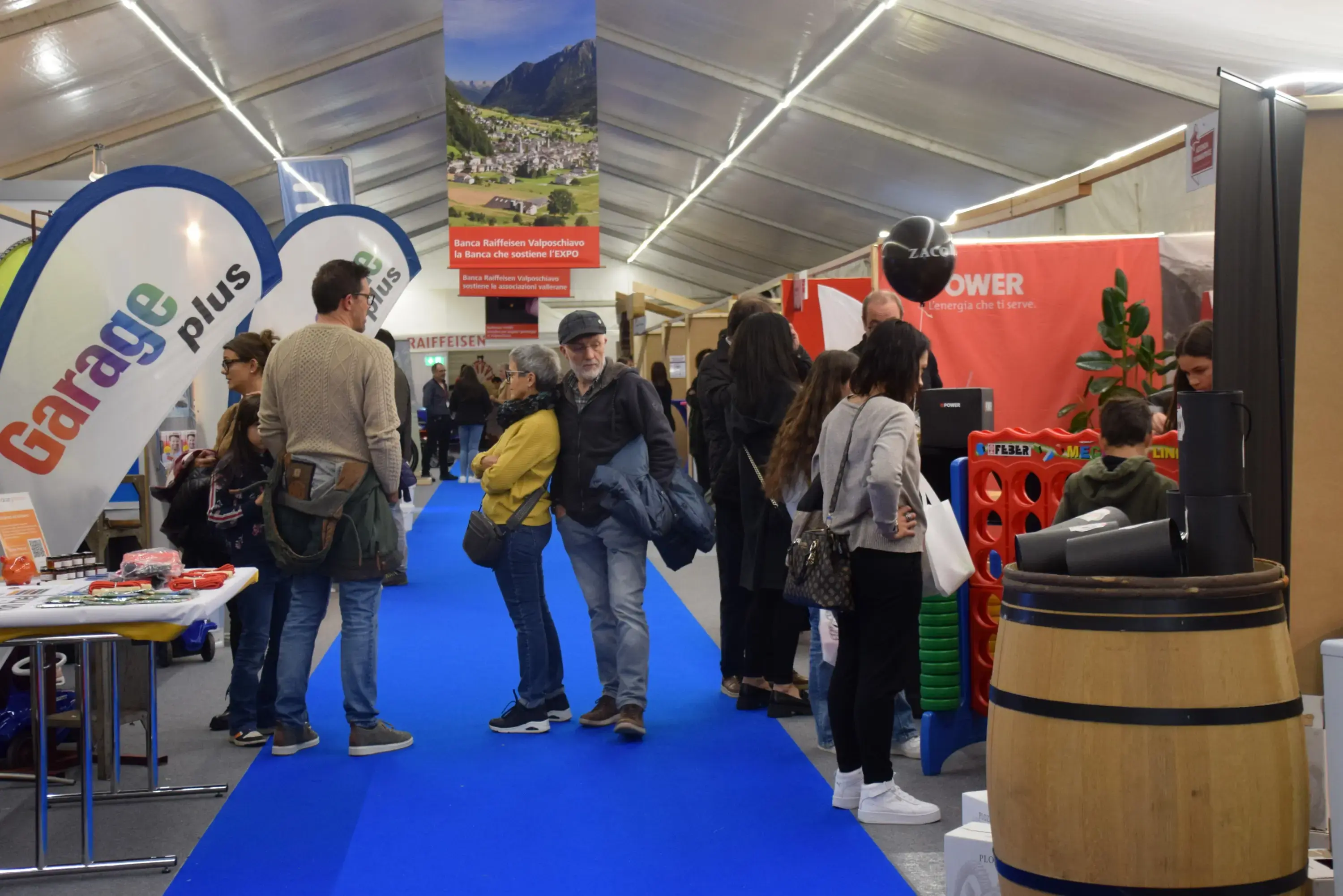 Un gruppo di persone si muove tra gli stand di una fiera, con espositori visibili e un pavimento blu. Si notano diverse bandiere e oggetti in mostra, mentre i visitatori interagiscono tra loro.