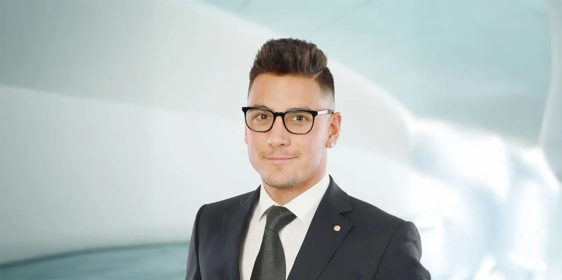 Uomo in abito elegante con cravatta e occhiali, sorriso fiducioso, su sfondo moderno e luminoso.