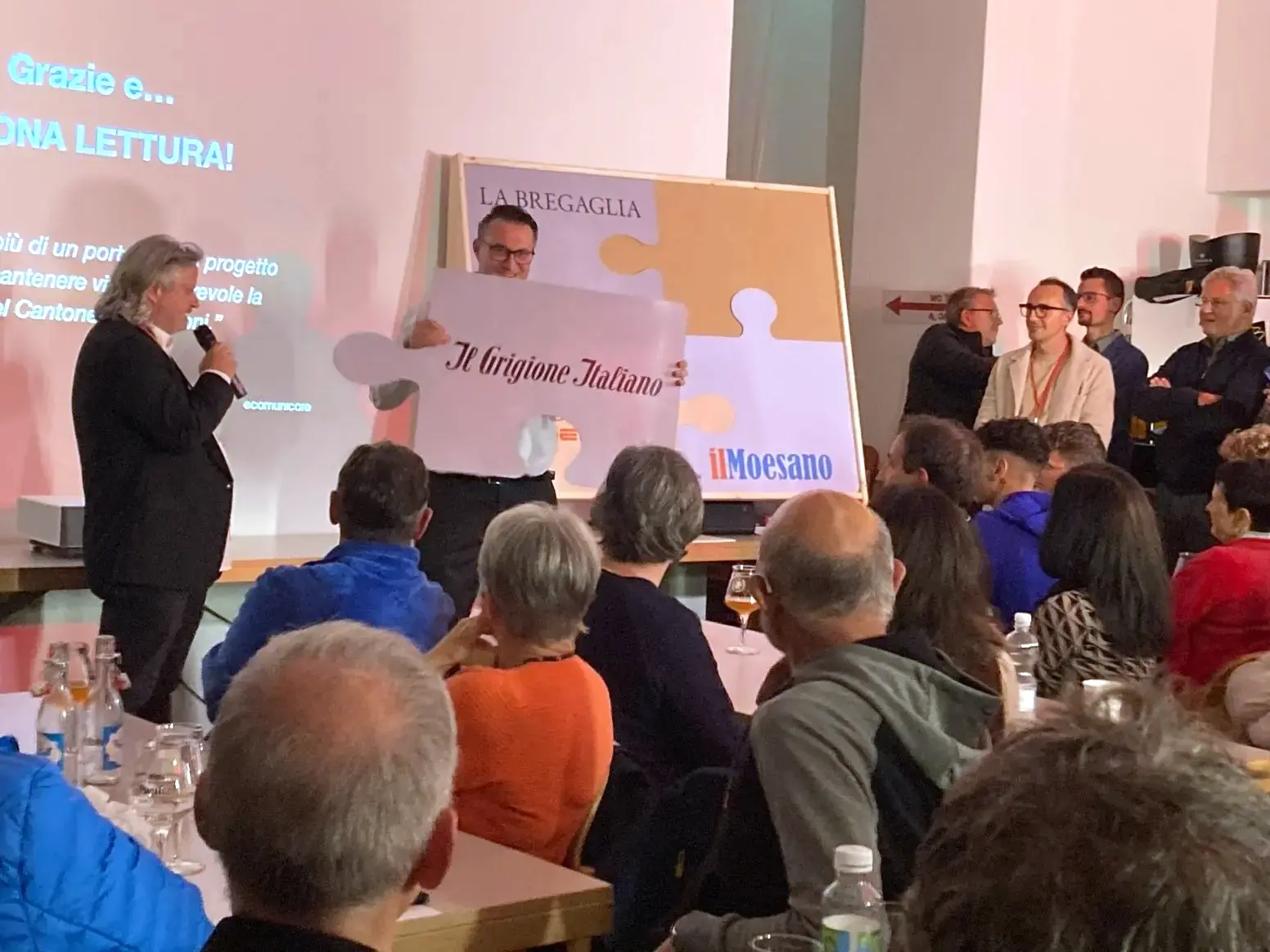Un gruppo di persone assiste a un evento, con un presentatore che mostra un grande cartello con la scritta "Il Grigionese Italiano". Sullo sfondo, una presentazione visiva e diversi partecipanti seduti a un tavolo.