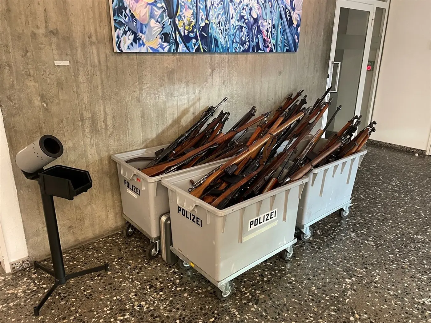 Fucili disposti in contenitori di plastica grigia etichettati "POLIZEI", posizionati su un pavimento in piastrelle di pietra. Sullo sfondo, si intravede una parete di cemento e un'opera d'arte.