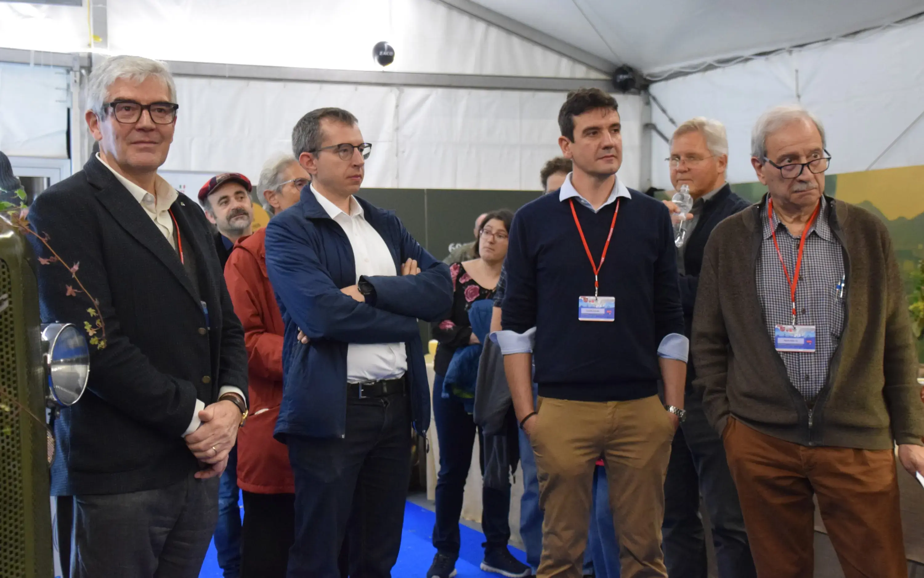 Gruppo di persone in un tendone, alcune in piedi in cerchio e altre sedute. Alcuni indossano occhiali e hanno un'espressione seria mentre osservano qualcosa di fronte a loro. Si notano curiosi dettagli come pass e badge appesi al collo.