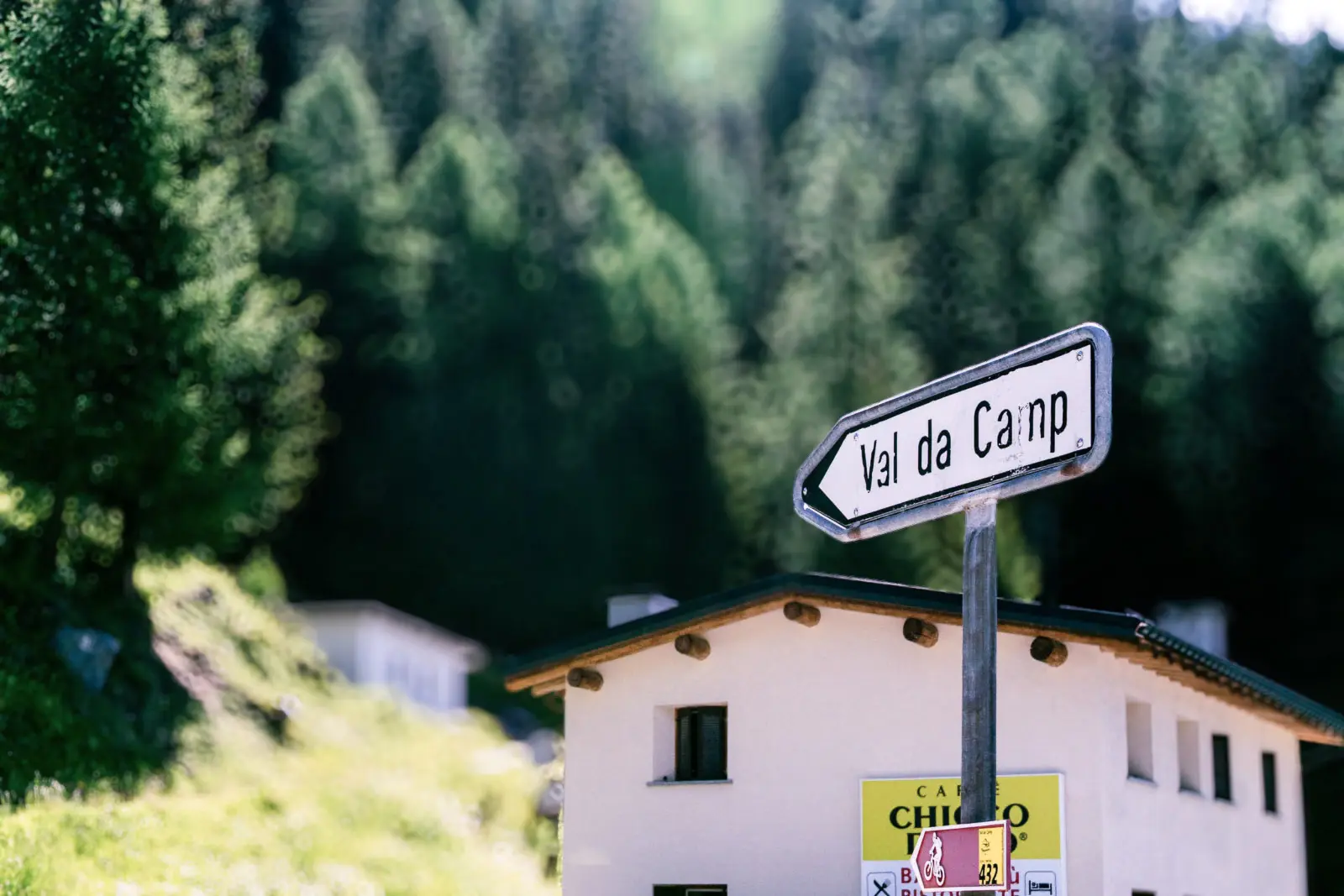 Insegna stradale che punta verso "Val da Camp", con un edificio in primo piano circondato da alberi e natura.