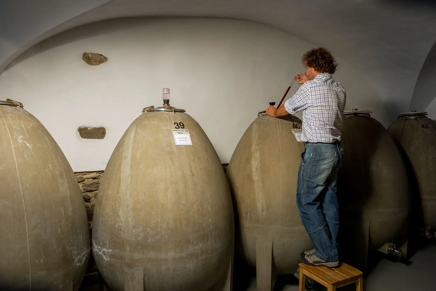 Un uomo in cima a una scala misura il contenuto di una grande botte di cemento a forma di uovo, all'interno di una cantina.