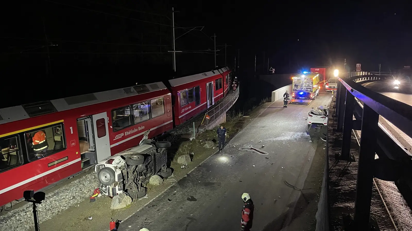Incidente ferroviario notturno con un treno deragliato su un binario; alcuni soccorritori sono in azione e un'auto ribaltata è visibile lungo la strada. Luci d'emergenza illuminano la scena.