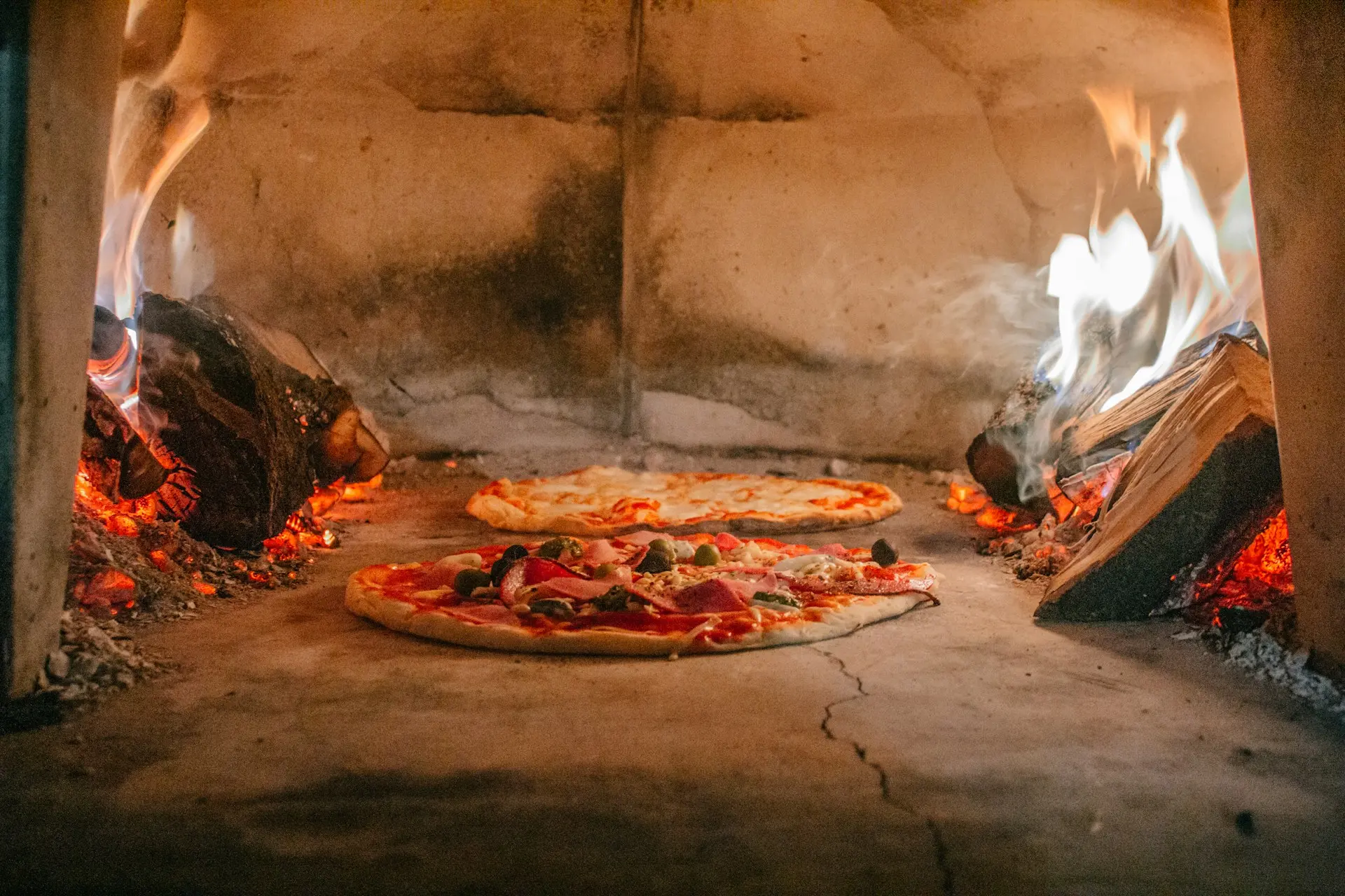 Due pizze in cottura in un forno a legna, con fiamme e braci visibili sullo sfondo.