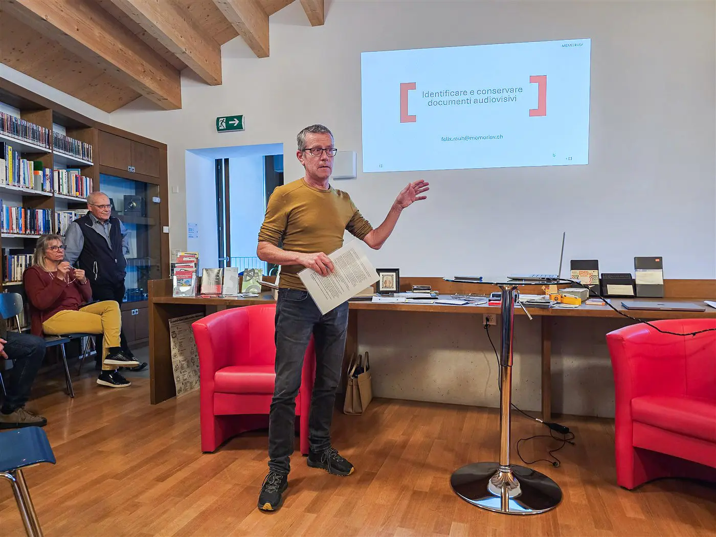 Un uomo in maglione giallo parla di fronte a un gruppo di persone sedute, mentre una presentazione è visibile su uno schermo. Il locale è una biblioteca con scaffali di libri in fondo.