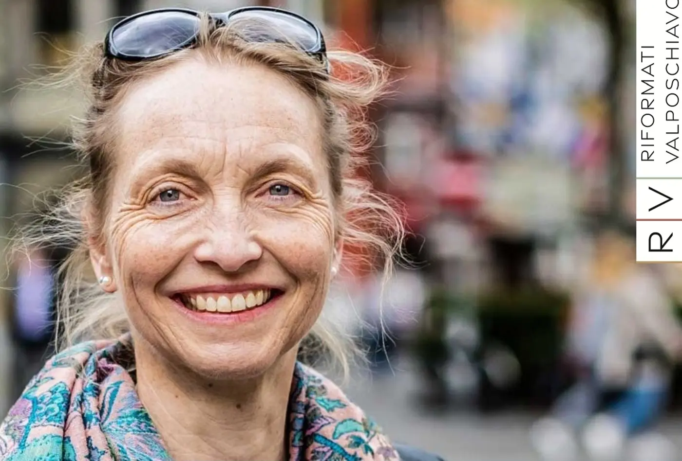 Una donna sorridente con capelli biondi raccolti e occhiali da sole, indossa una sciarpa colorata. Sullo sfondo, si intravedono persone e un ambiente urbano sfocato.