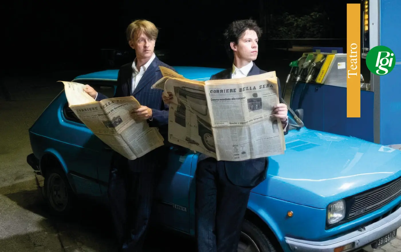 Due uomini in abiti eleganti leggono dei giornali mentre sono appoggiati a un'auto blu in un'area di servizio. Una luce notturna illumina la scena, creando un'atmosfera interessante.