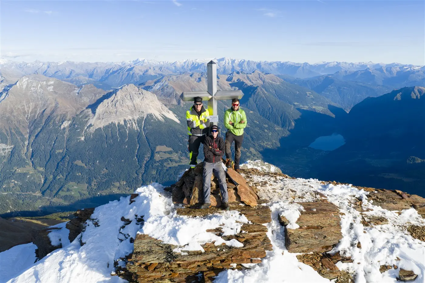 Due uomini in giacca alta visibilità e un uomo con giacca scura stanno in piedi accanto a una grande croce di metallo sulla cima di una montagna. Sullo sfondo si vedono montagne innevate e un lago in lontananza.