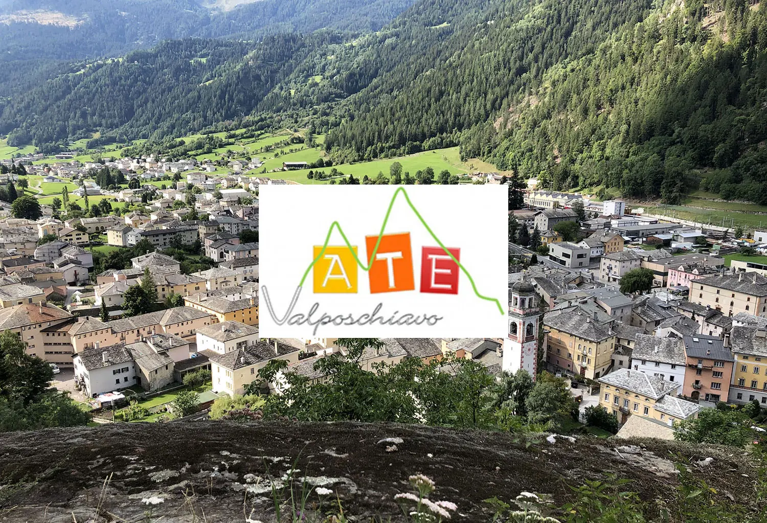 Panorama di un paese circondato da monti verdeggianti, con case tradizionali e un cielo parzialmente nuvoloso. Al centro, si trova il logo colorato "ATE Valposchiavo".