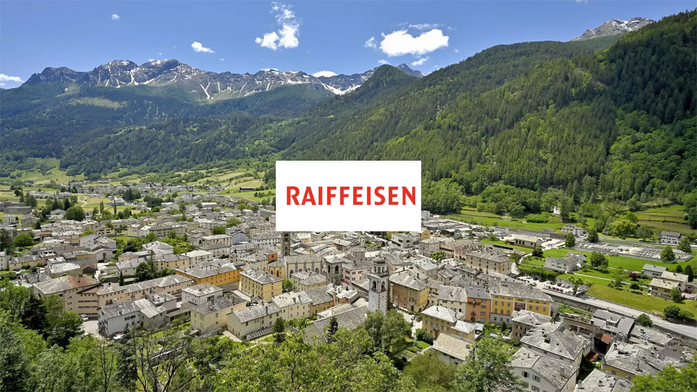 Panorama di un villaggio montano con case dai tetti di tegole, circondato da montagne verdi e innevate. In primo piano si trova il logo "Raiffeisen" in rosso su sfondo bianco.