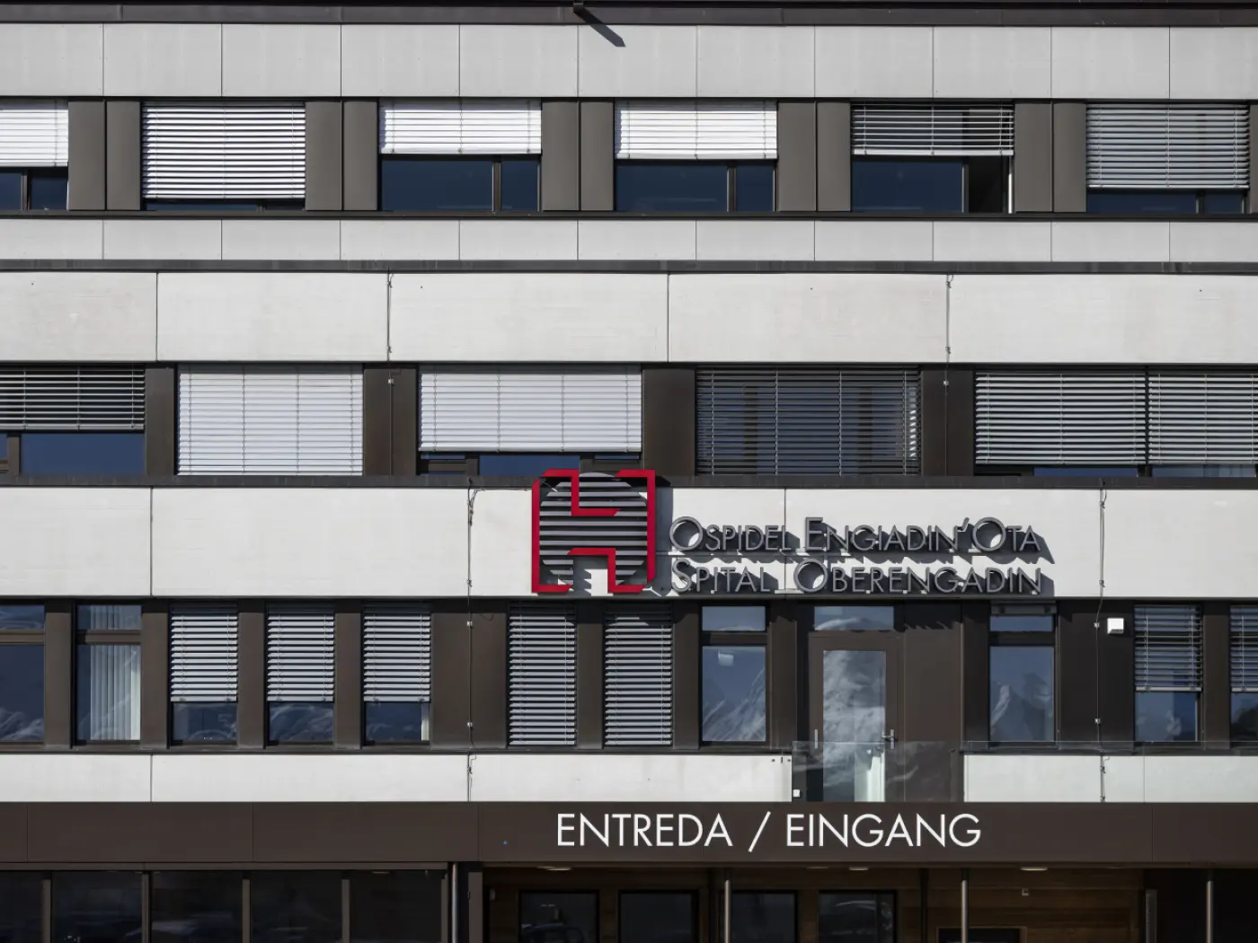 Facciata di un ospedale con un'insegna che indica "Ospidal Engiadina" e le parole "Entrata / Eingang" in diverse lingue. Le finestre hanno persiane chiuse.