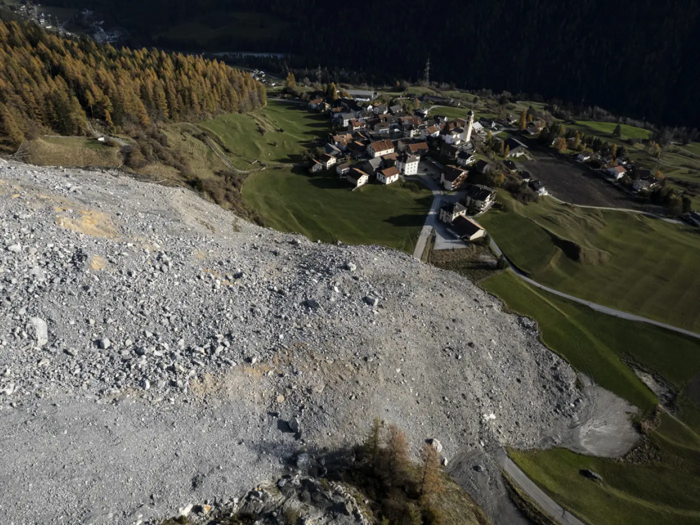 Un'area di frana con rocce e detriti che scendono verso un villaggio rurale circondato da alberi di conifere e campi verdi. La chiesa del villaggio è visibile tra le abitazioni.