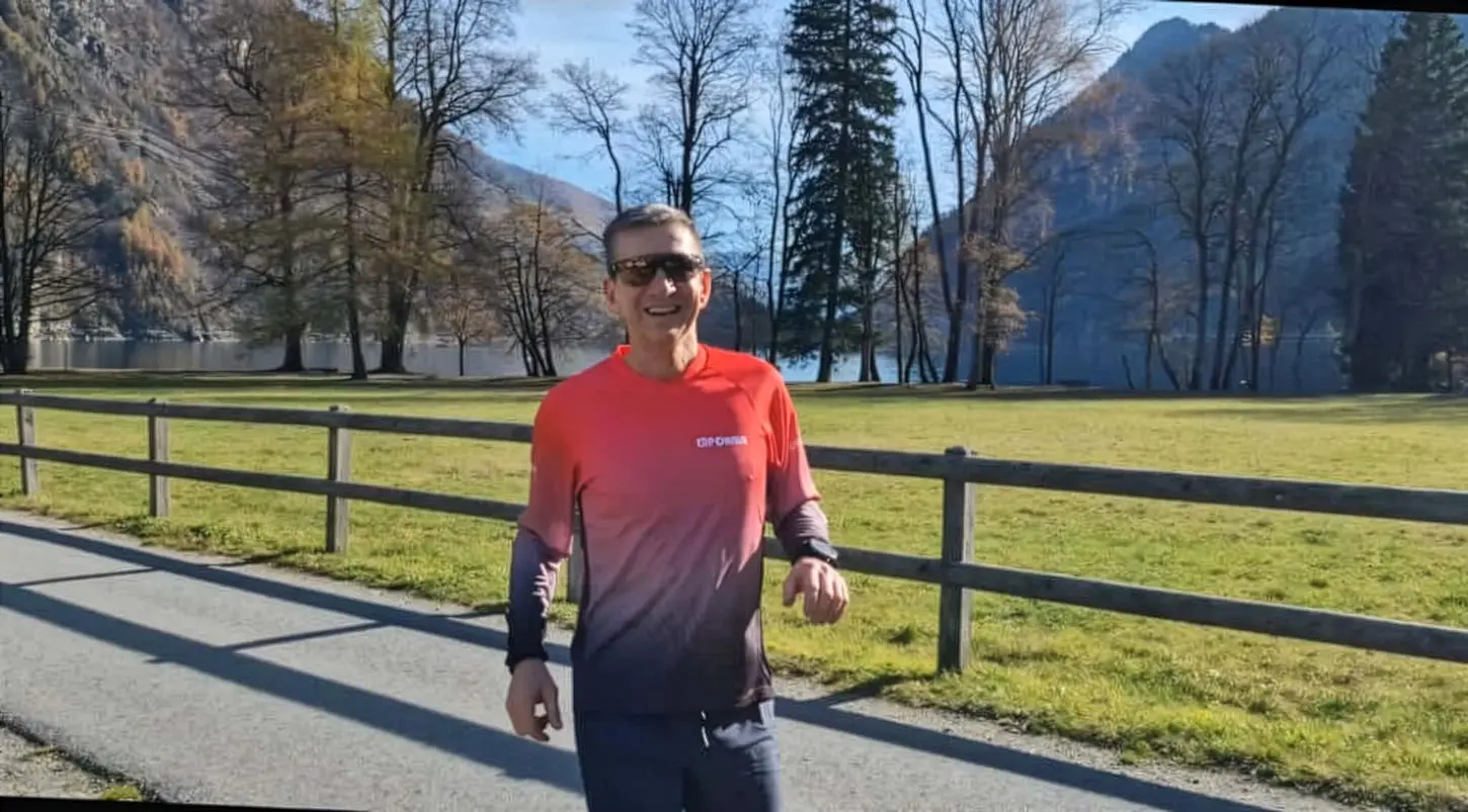 Un uomo che corre su un sentiero nel parco, circondato da alberi e montagne. Indossa una maglietta rossa e grigia e sorride mentre si allena.