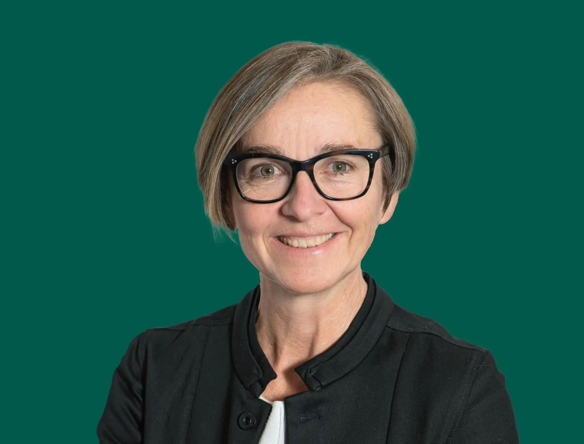 Donna sorridente con occhiali neri e capelli corti e grigi, indossa una giacca nera su sfondo verde.